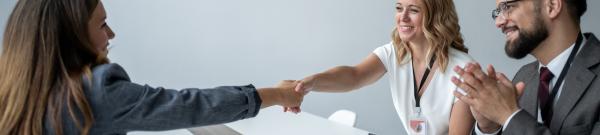 Two professionally dressed women shaking hands next to a professionally dressed man clapping