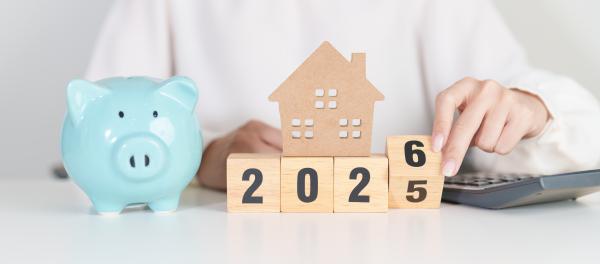 Wooden blocks reading 2025 with a woman sat behind turning the "5" over to become a "6". Blue piggy bank on one side of the blocks, calculator on the other
