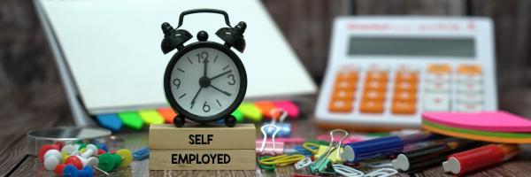 Image of a busy desk with a small alarm clock on top of two wooden blocks with self-employed on them, and a calculator and pad in the background