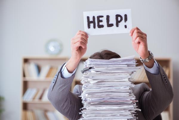 A man hidden behind a pile of papers holding up a 'help' sign