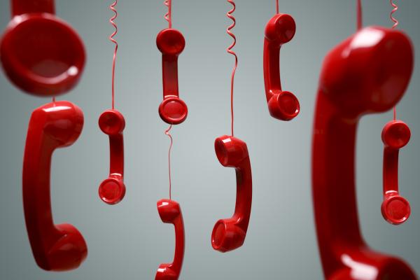 A number of red old-fashioned phone handsets are dangling by their wires against a grey backdrop