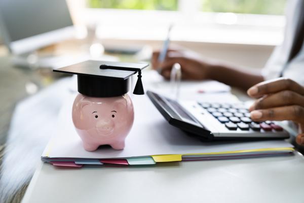 Person at desk using calculator and doing paperwork. A piggy bank wearing a graduate's mortar board sits on the desk.