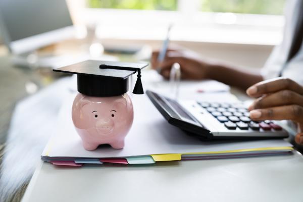 Person at desk using calculator and doing paperwork. A piggy bank wearing a graduate's mortar board sits on the desk.