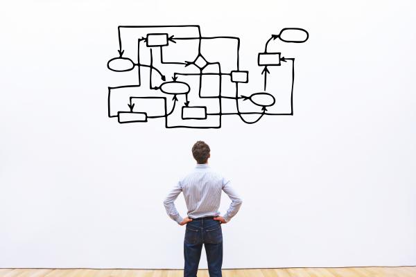 A man is staring at a white wall with a complex diagram of boxes all linked together in a confusing pattern. The man has his back to us and is wearing blue jeans with his hands on his hips. 