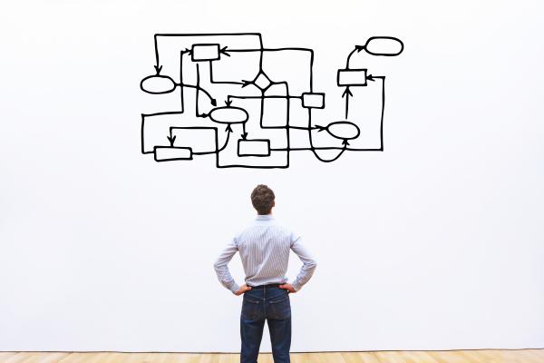 A man is staring at a white wall with a complex diagram of boxes all linked together in a confusing pattern. The man has his back to us and is wearing blue jeans with his hands on his hips.