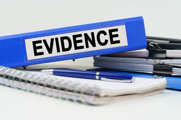 A blue folder with a white label saying 'evidence' sits on top of a pile of pens and ring-binder note books 