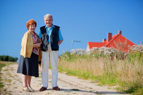 A elderly couple stand outside their property. The house has a red roof and is hidden by long grasses, suggesting it might be beside the sea