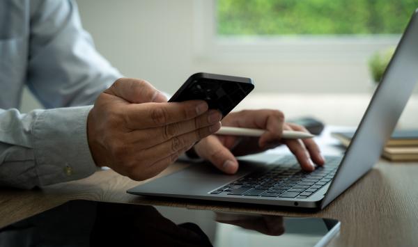 Laptop on desk with man typing whilst using mobile phone with his other hand