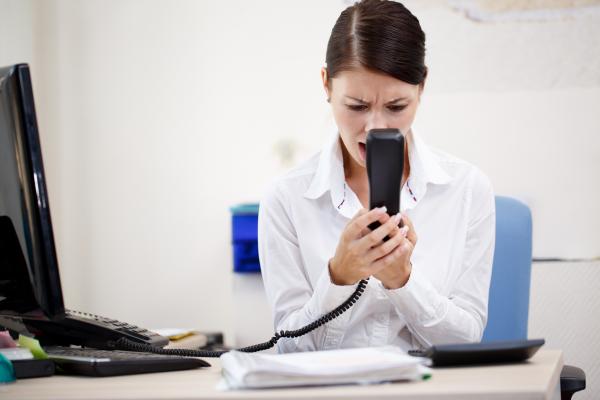 A women wearing office attire is sitting at her laptop, holding a phone in her hands and shouting at it.