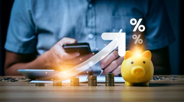 Piles of coins of increasing sized on desk next to a piggy bank with virtual percentage sign and arrow tracking increase