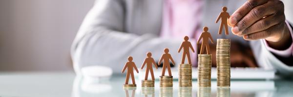 Piles of coins of increasing sized on desk with cardboard people figures on top of each coin