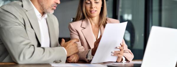 Man and woman sat together referring to piece of paper with laptop on table infront of them