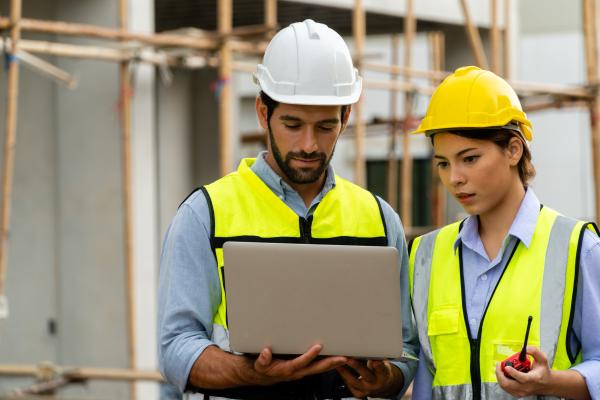 Man and woman in yellow hi-viz vests and hard hats using a laptop, with construction site in the background
