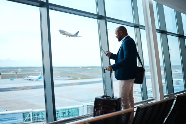 Man stood at airport window with a suitcase, using mobile phone. Plane taking off in the background