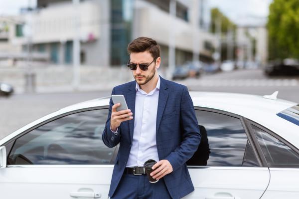 Image shows a man in suit leaning on white car and using a mobile phoone