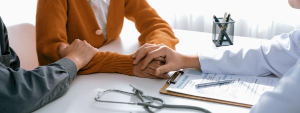 Doctor sat opposite a couple in a medical consulting room, holding the woman's hands on the desk while her partner holds her arm