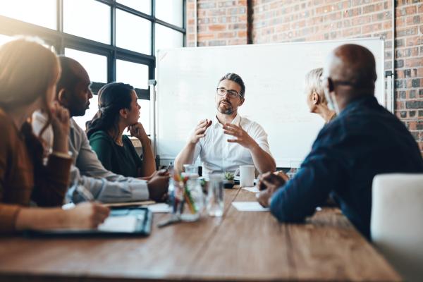 Five people sat around a rectangular desk in a meeting room, listening to man at the end presenting 