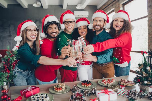 Group of people with Santa hats on stood behind table of buffet food and clinking glasses. Christmas decorations in the background