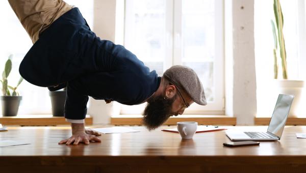 Man doing inverted yoga pose on a desk next to coffee cup and laptop
