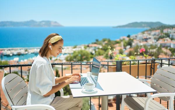 Woman using laptop on a balcony in sunny location with sea and Mediterranean scene in the background