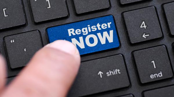 A finger is pressing a blue button on a keyboard which reads 'register now'. 