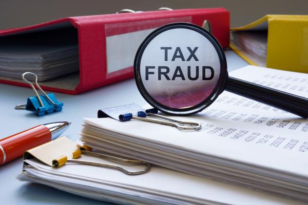 Piles of papers on a desk, magnifying glass resting on its side on the top and magnifying "Tax Fraud" written on a folder in the background