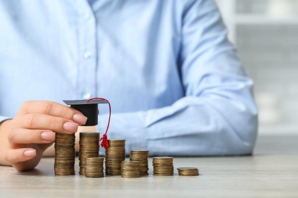 Stacks of coins in a row on a desk, with a hand holding a university-style mortar board hat over the larger stacks