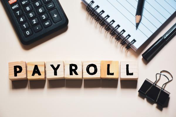 Wooden blocks reading "Payroll" on a desk, surrounded by calculator, notepad and bulldog clip