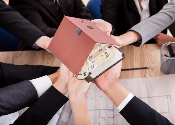 Multiple hands reaching out over a table to jointly support a model house