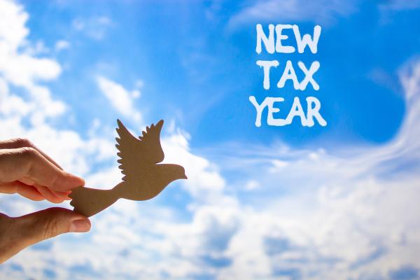 "New Tax Year" written in white against a blue sky with white clouds, a hand holds a cardboard cutout of a bird flying on the left of the image