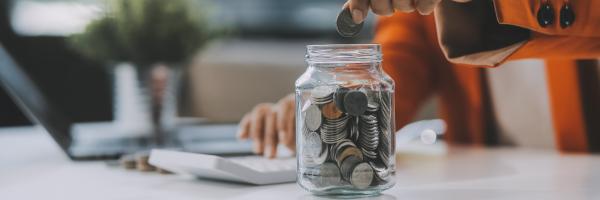 Image shows a hand dropping a coin into a jar of money, with a calculator in the background