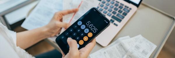 Image shows someone adding up receipts on a phone calculator with a laptop in the background