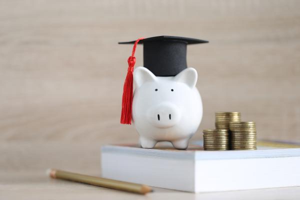 White piggy bank wearing a mortar-board hat sat on a textbook next to a pile of coins