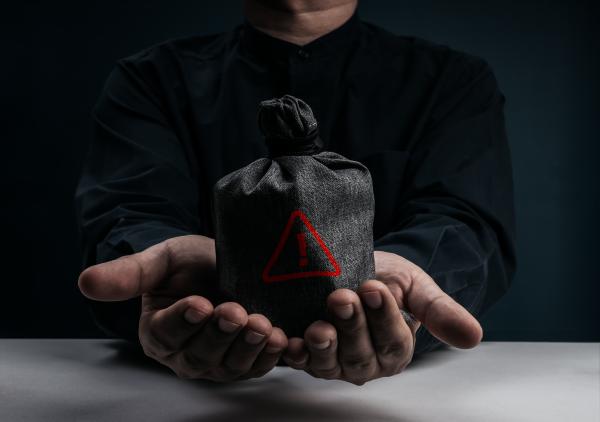 Dark image of man dressed in dark clothes holding a grey money bag in his outstretched hands with a red exclamation mark on it 