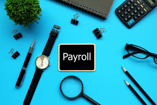 "Payroll" written on a small blackboard surrounded by desktop items (pen, glasses, laptop, magnifying glass etc)