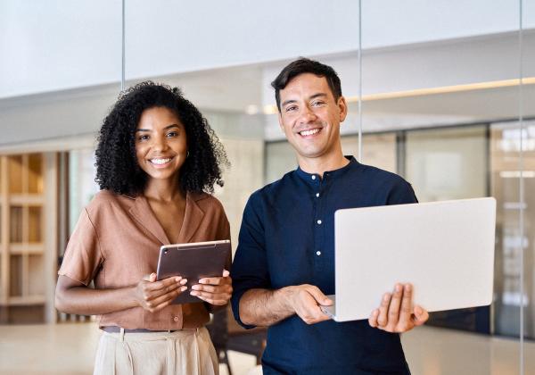 Woman and man stood in a professional setting holding laptop/tablet and smiling