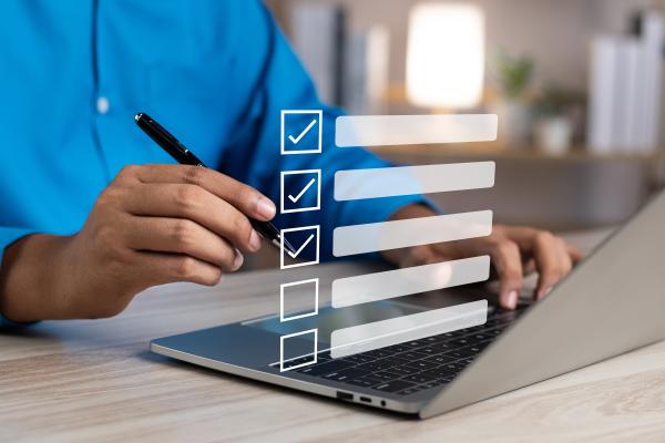 A man in a blue shirt is sitting at a laptop, ticking off items on a list
