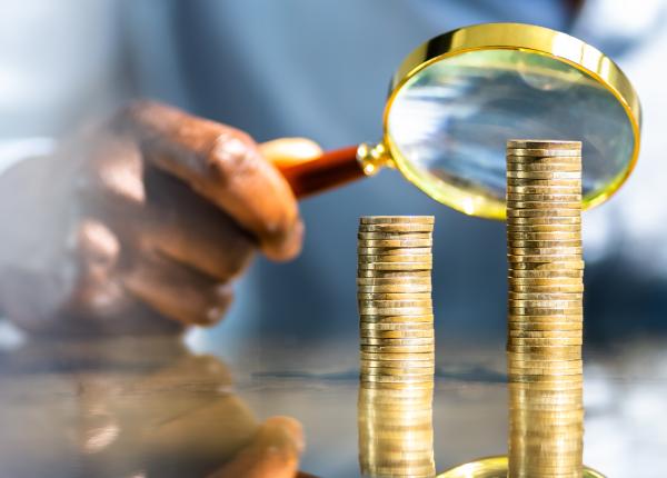Hand holding magnifying glass over two piles of coins, one of which is taller than the other