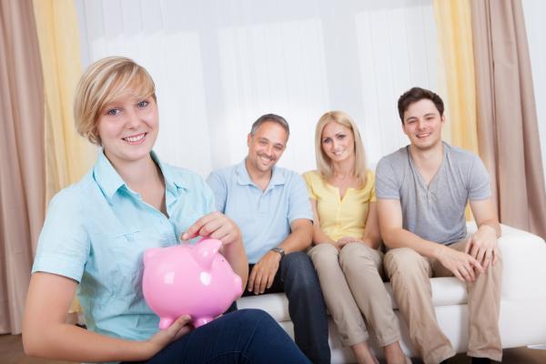 Two adults sat on sofa next to a teenage boy, with teenage girl in foreground  putting coin in a piggy bank